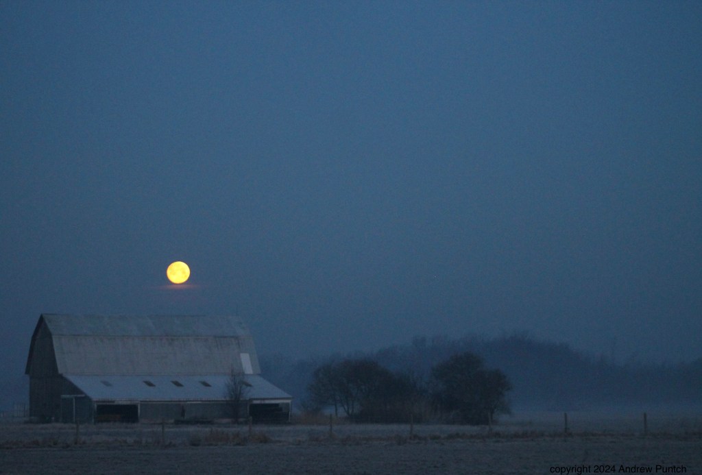 A metal barn softly illuminated by morning light, with the full or nearly full moon setting above it.